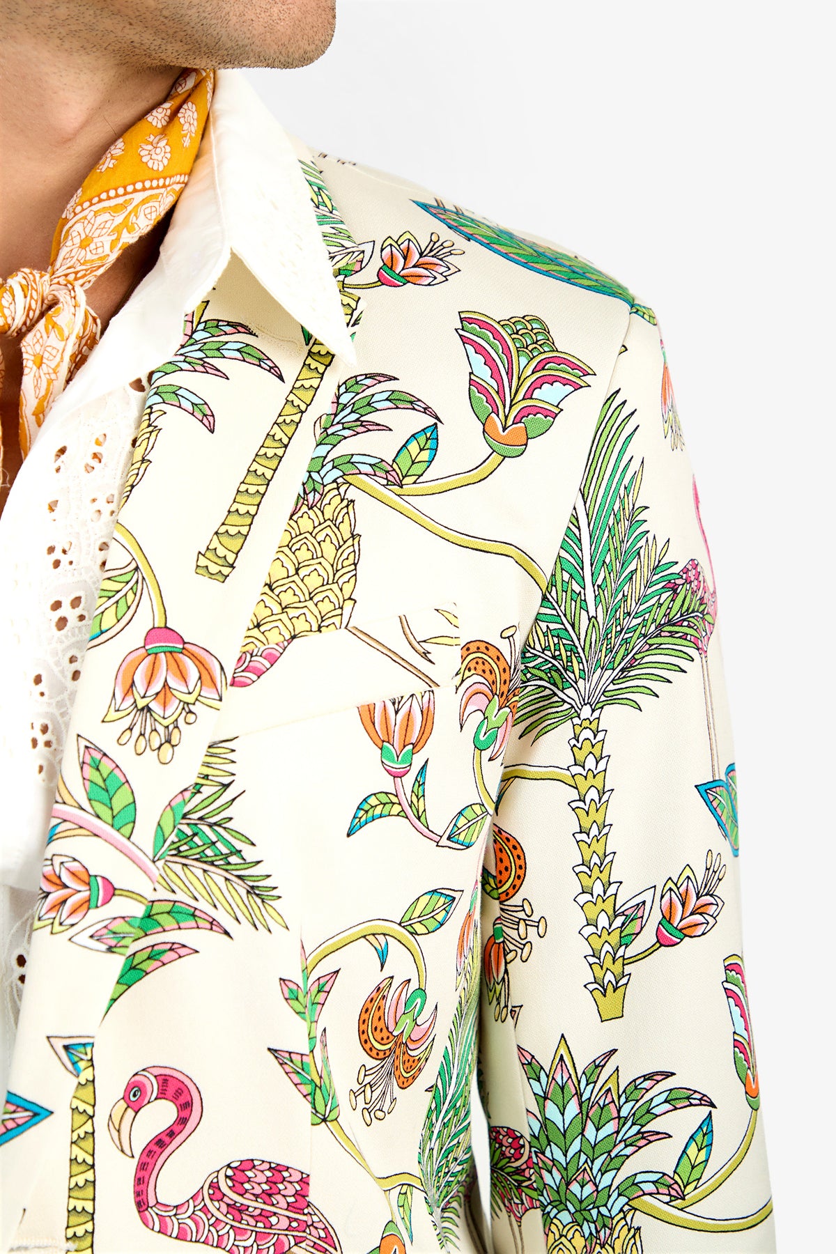 A close-up of a person wearing the THURSTON BLAZER, a cream-colored cotton blazer with a vibrant tropical print of palm trees, flowers, and a pink flamingo. They pair it with a white shirt and a patterned yellow lycra scarf.
