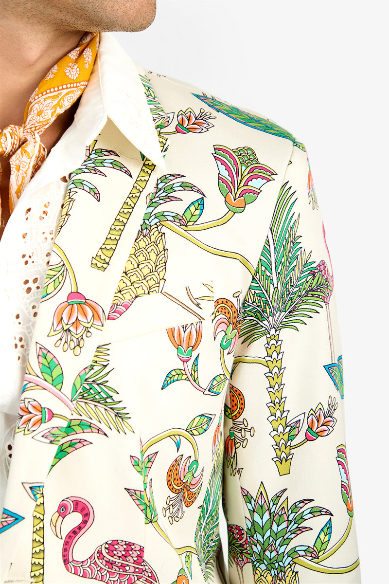 A close-up of a person wearing the THURSTON BLAZER, a cream-colored cotton blazer with a vibrant tropical print of palm trees, flowers, and a pink flamingo. They pair it with a white shirt and a patterned yellow lycra scarf.