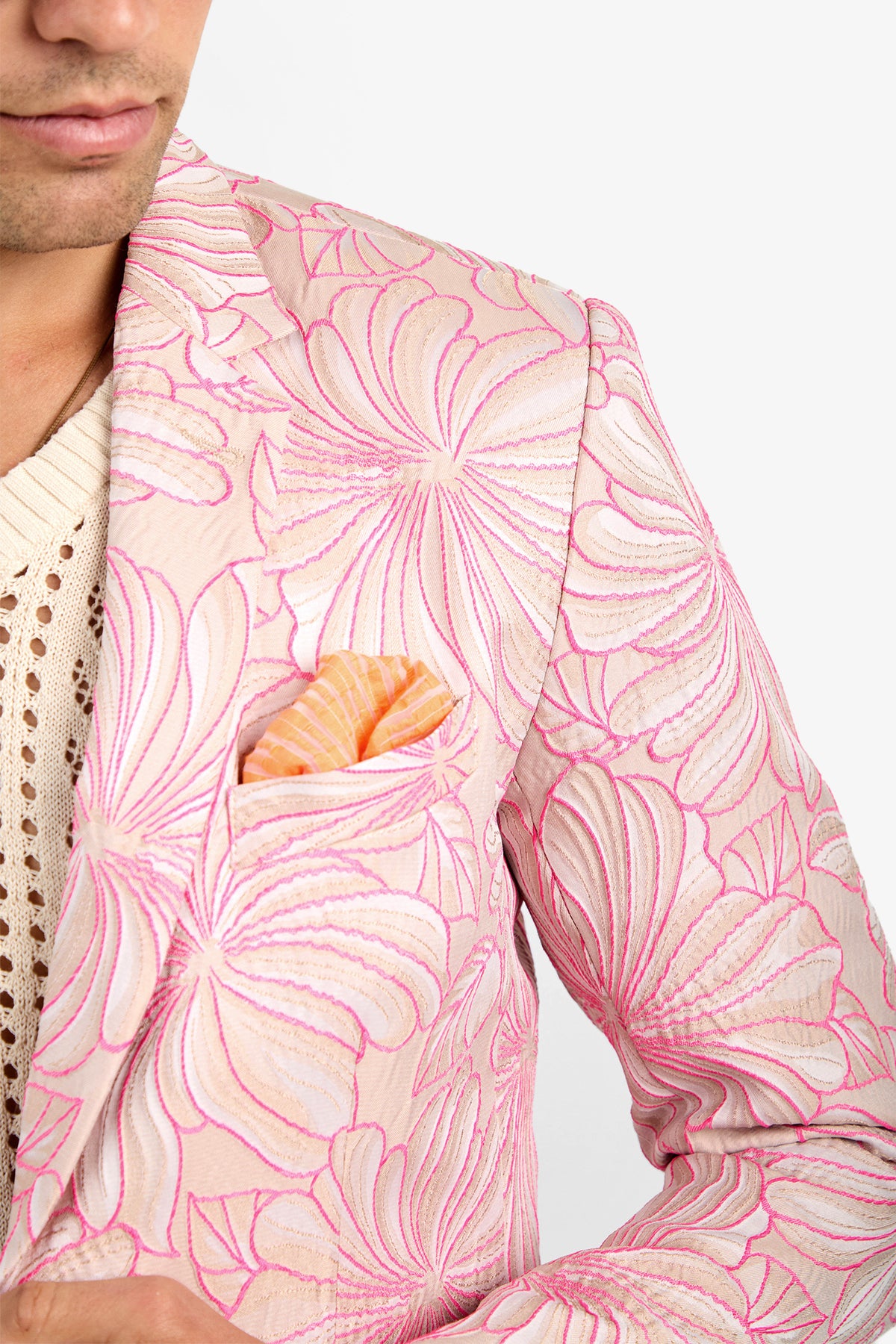 A person wearing the THURSTON BLAZER—a beige jacket with bold pink floral embroidery—over a cream knit top and an orange-striped pocket square. Only their lower face and upper body are shown.