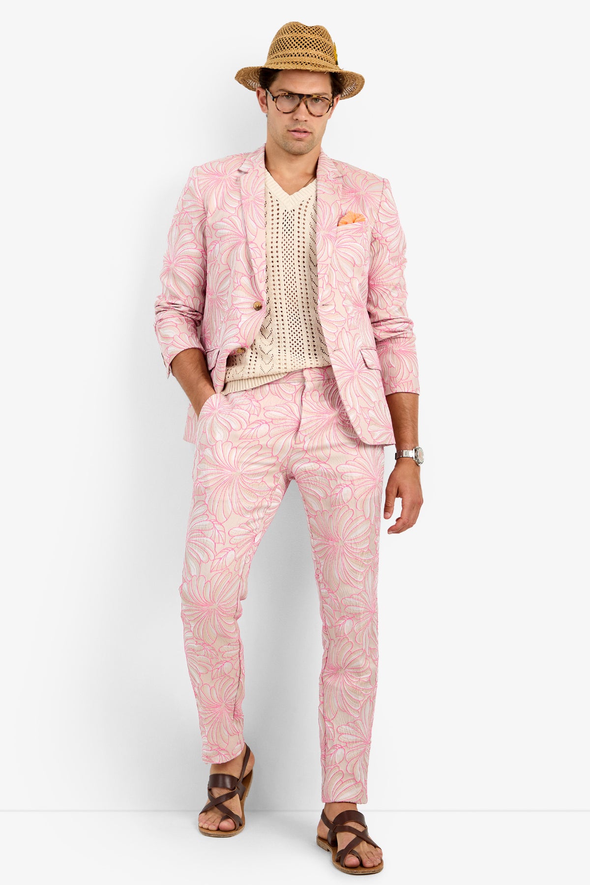 A man confidently poses in CLYDE SLIM TROUSER, a light pink cotton suit with a floral pattern, paired with a cream crochet shirt, brown sandals, a straw hat, glasses, and a watch against a white background.