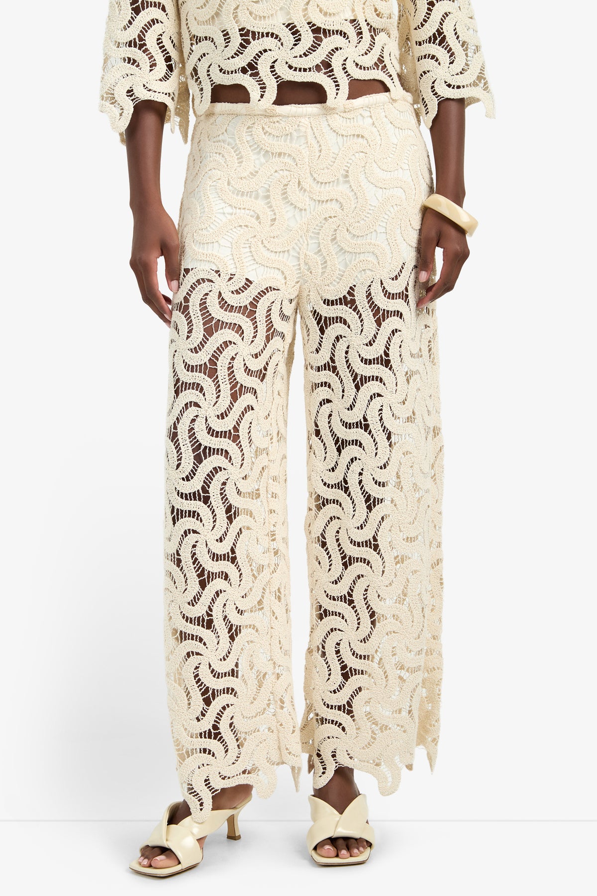 A person wears the BARDAN PANT—wide-leg, cream crochet lace pants made from 100% cotton with a wavy pattern—styled with a matching top, cream sandals, and a chunky bracelet against a plain white background.