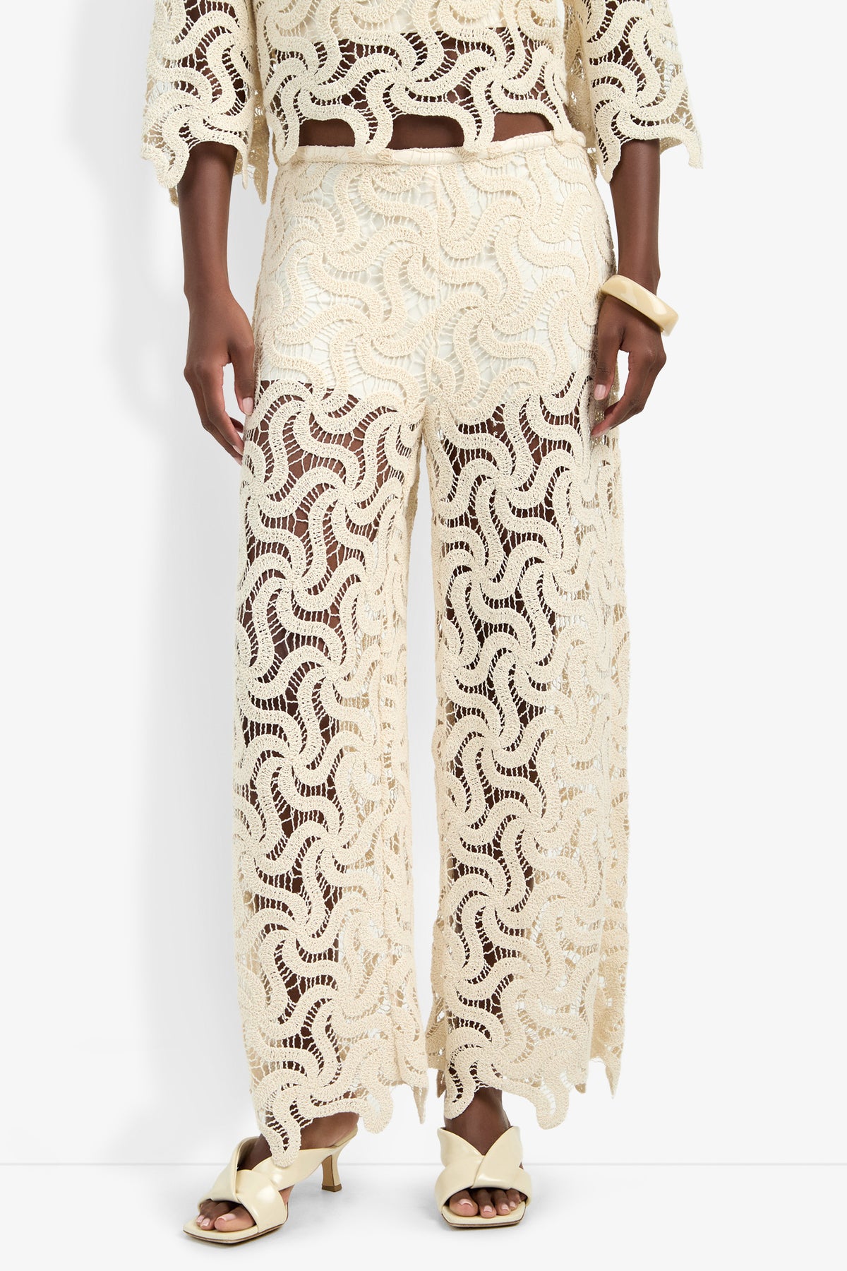 A person wears the Bardan Pant in cream, featuring sheer lace and an intricate wavy pattern. Matching lace top partially visible, styled with cream sandals and a chunky bracelet.