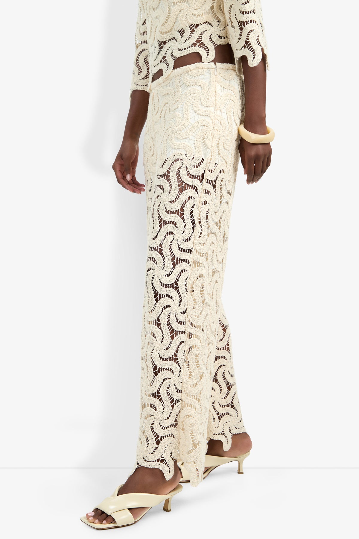 A person wears the Bardan Pant, a cream-colored, sheer crocheted style paired with a matching top, cream heeled sandals, and a chunky cream bracelet against a white background.