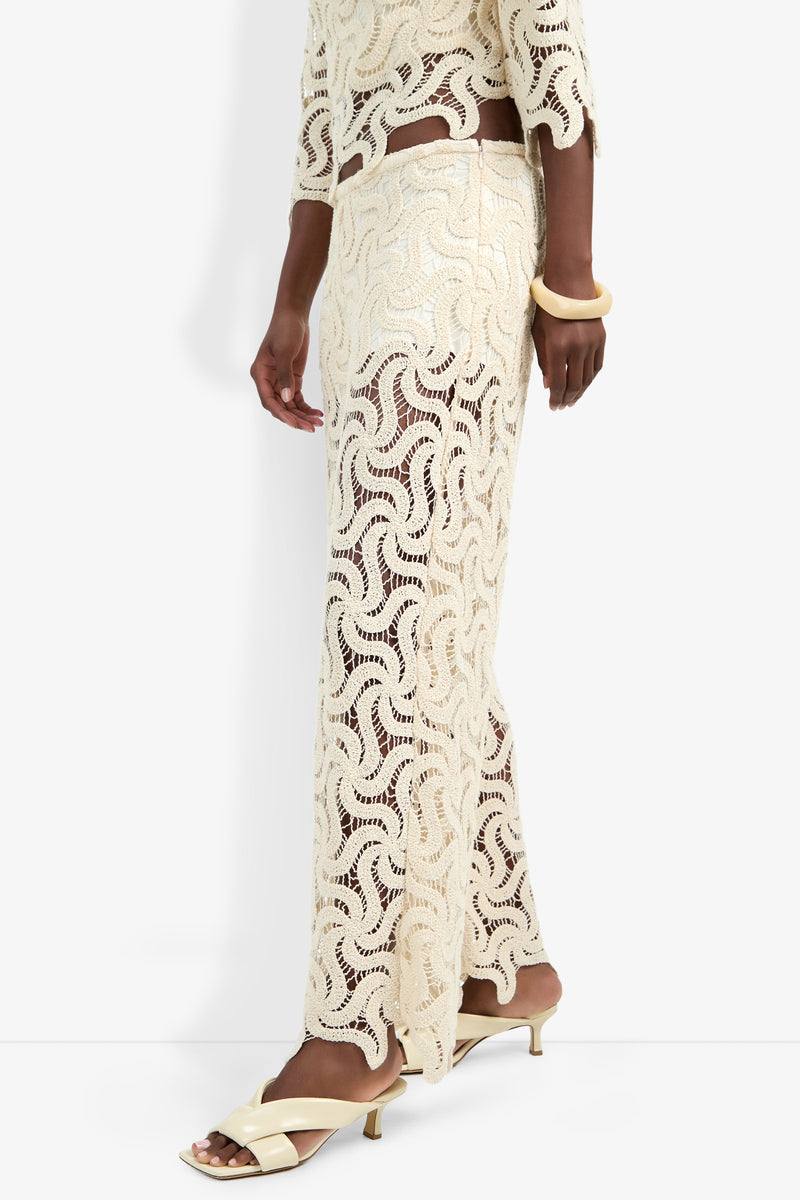 A person wears the Bardan Pant, a cream-colored, sheer crocheted style paired with a matching top, cream heeled sandals, and a chunky cream bracelet against a white background.