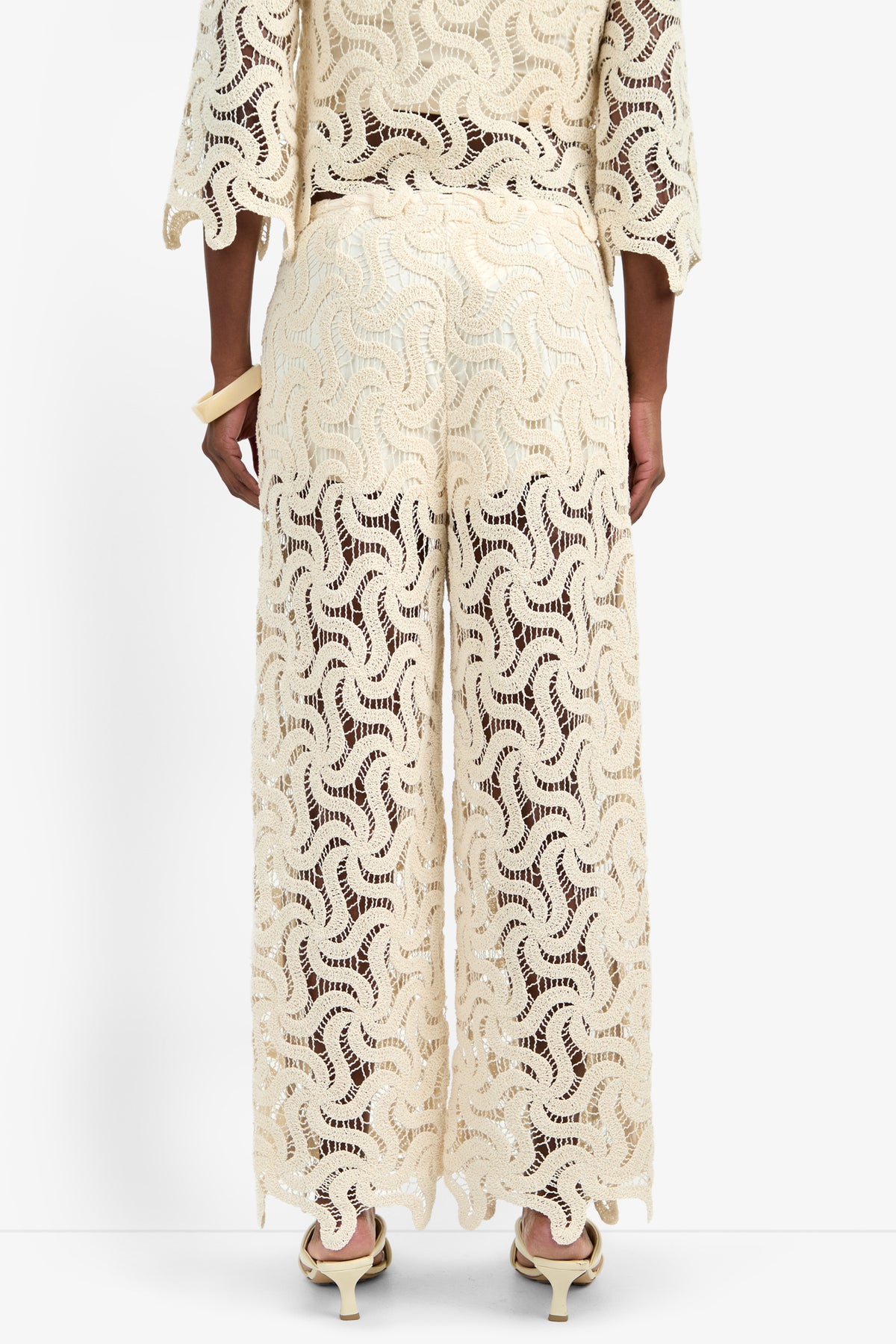 A person stands facing away, wearing the BARDAN PANT—cream-colored, wide-leg cotton pants with a wavy lace pattern. Semi-sheer and stylish, they're paired with a matching top and cream high-heeled sandals.