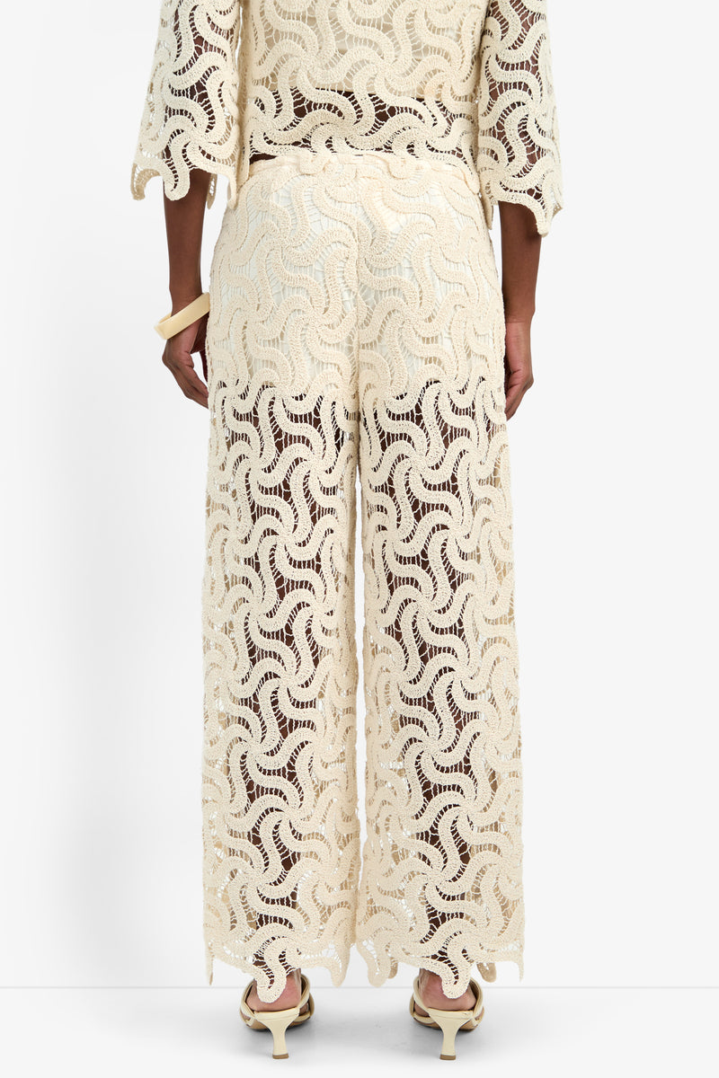A person stands facing away, wearing the BARDAN PANT—cream-colored, wide-leg cotton pants with a wavy lace pattern. Semi-sheer and stylish, they're paired with a matching top and cream high-heeled sandals.