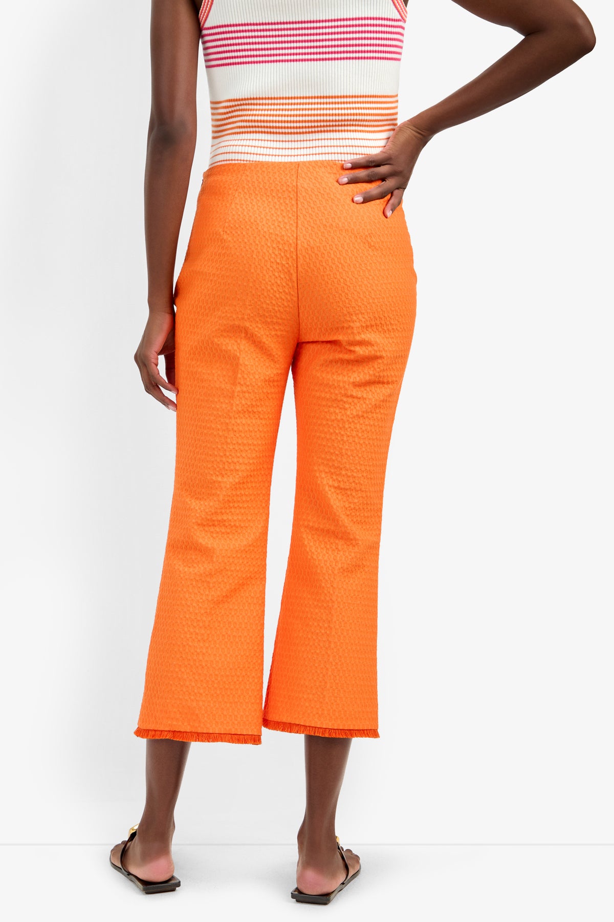 Wearing the GULF PANT in bright orange polyamide-elastane, paired with a sleeveless striped top and flat sandals, a person stands with one hand on their hip, seen from the back.