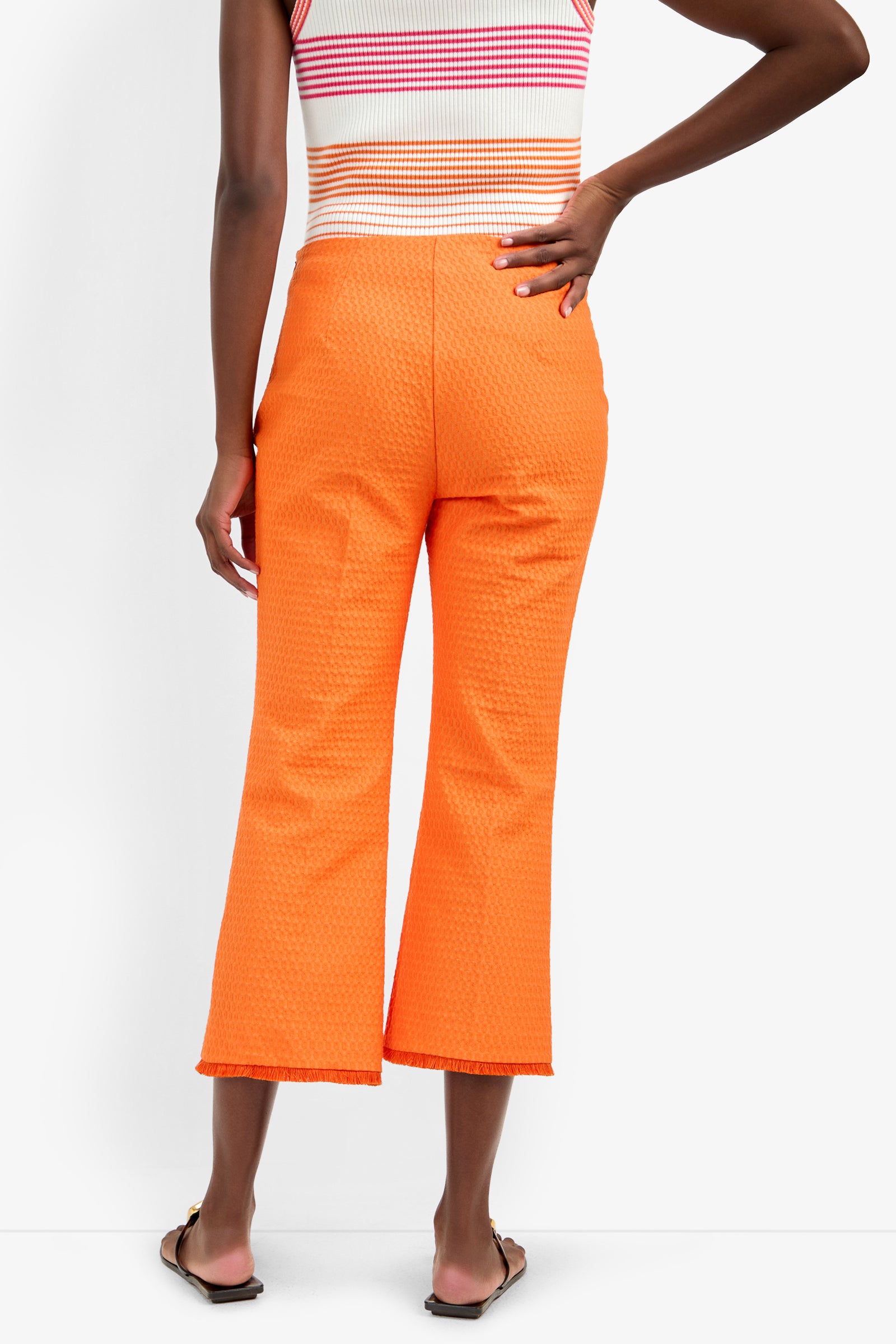Wearing the GULF PANT in bright orange polyamide-elastane, paired with a sleeveless striped top and flat sandals, a person stands with one hand on their hip, seen from the back.