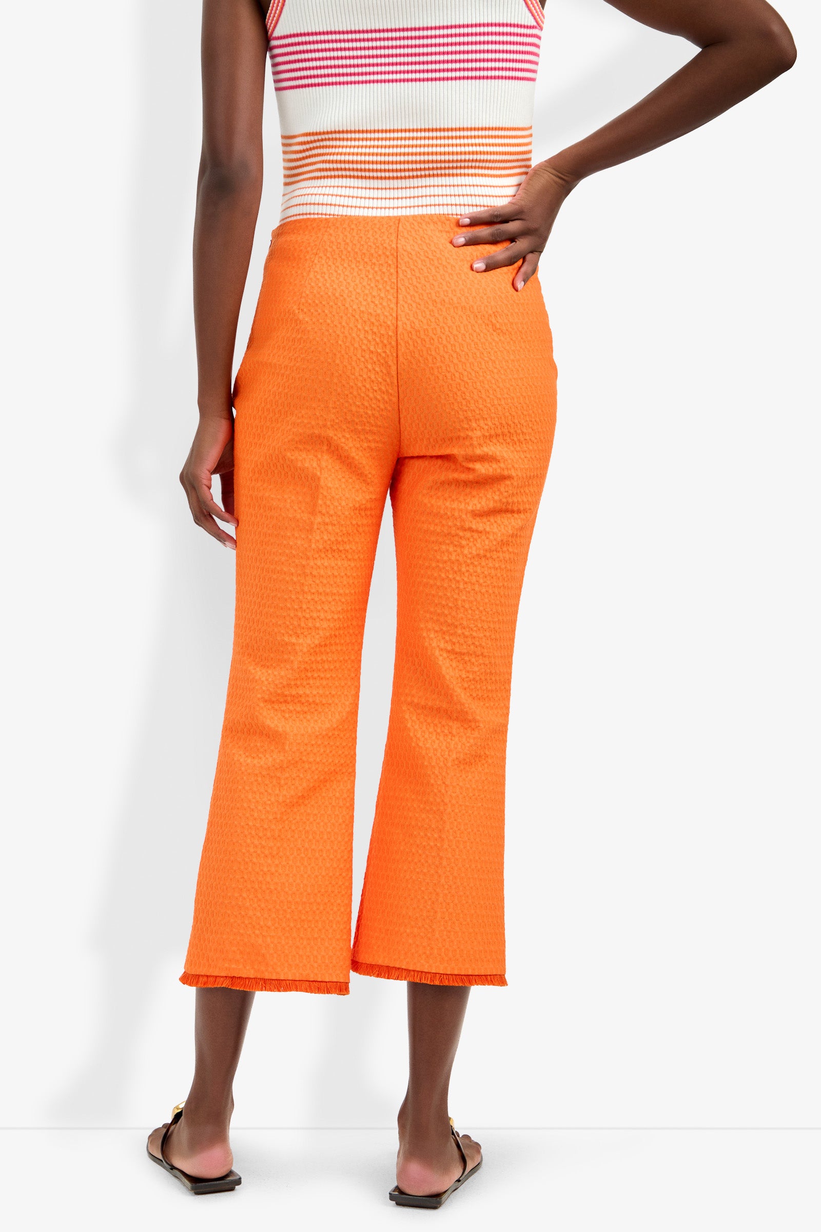 A person seen from the back wears bright orange Gulf Pant with a textured finish and a sleeveless striped top, standing with one hand on their hip and the other by their side.