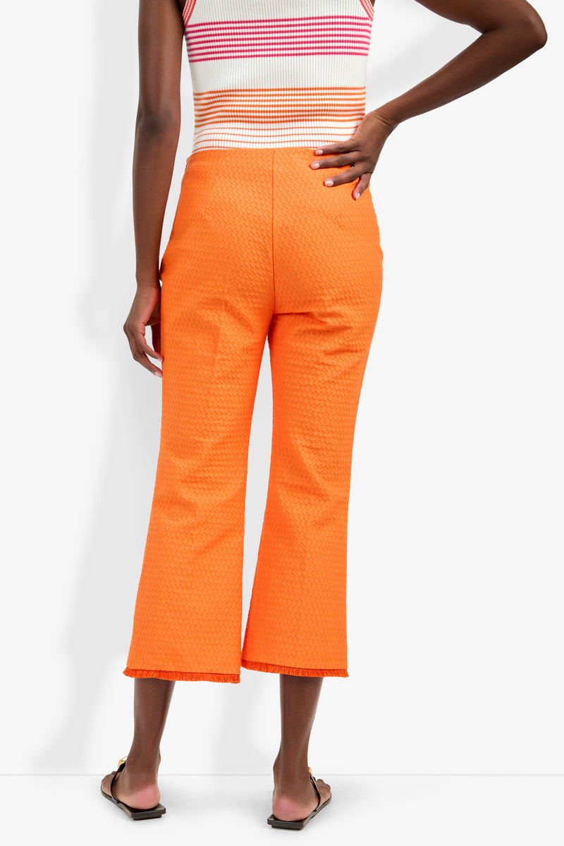 A person seen from the back wears bright orange Gulf Pant with a textured finish and a sleeveless striped top, standing with one hand on their hip and the other by their side.
