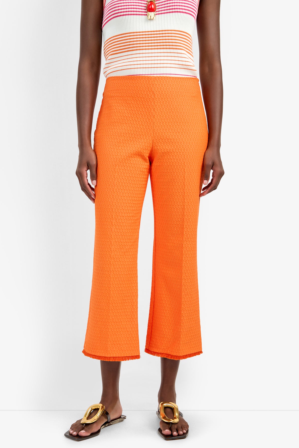 A person wears the GULF PANT in bright orange viscose blend with a textured, cropped fit, paired with a striped sleeveless top and brown flat sandals with gold buckles, standing against a plain background.