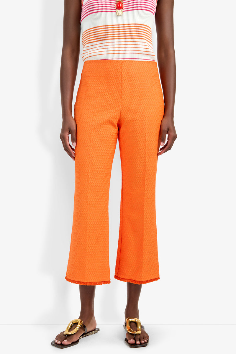 A person wearing Gulf Pant in bright orange with a textured, cropped design, paired with a colorful striped sleeveless top and brown sandals featuring gold embellishments, stands against a white background.