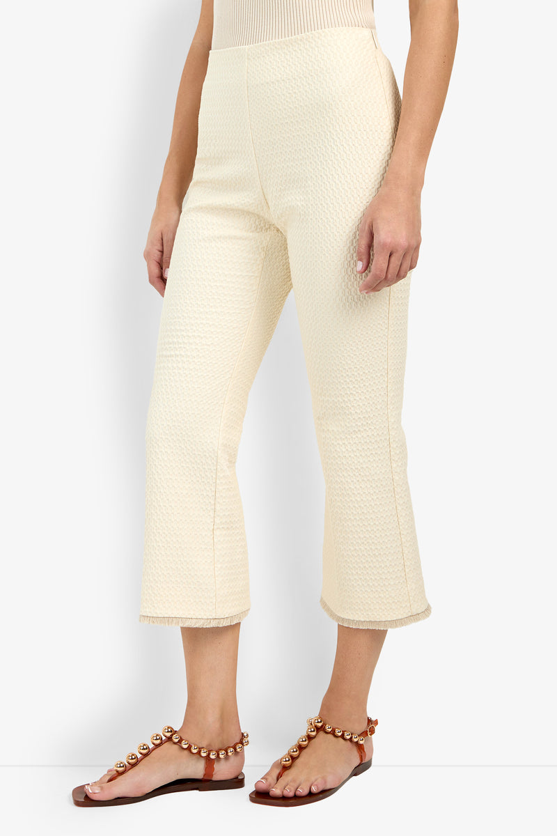 A person wears Gulf Pant—textured, cream-colored cropped pants—paired with brown flat sandals featuring decorative pearls, standing against a plain white background.