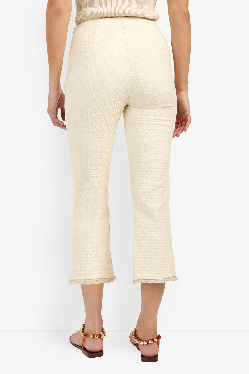 A person stands facing away, wearing the cream textured Gulf Pant—high-waisted with slight ankle flares—and a sleeveless top. They also wear brown sandals featuring gold chain details.