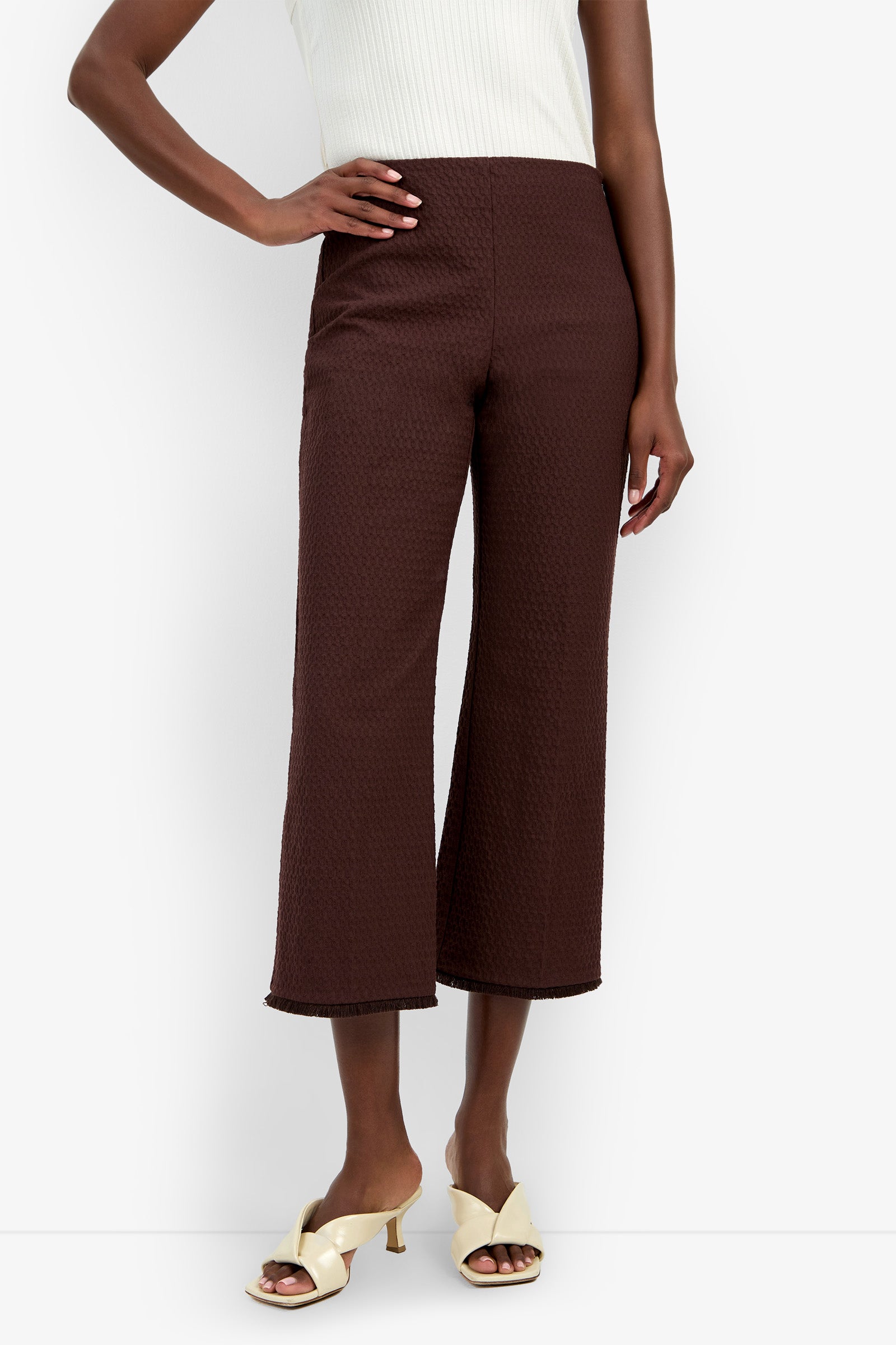 A person wearing the Gulf Pant—a pair of high-waisted, brown, cropped wide-leg pants—along with a white sleeveless top and cream heeled sandals, standing against a white background.