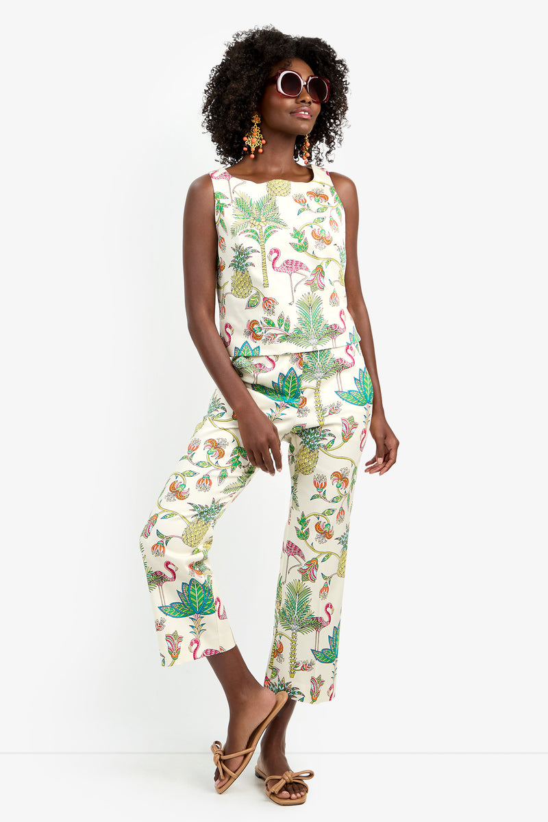 A woman models LULU PANT, a matching set of sleeveless, tropical print cotton top and spandex pants with birds and plants. She accessorizes with round sunglasses, colorful earrings, and brown sandals against a plain white backdrop.