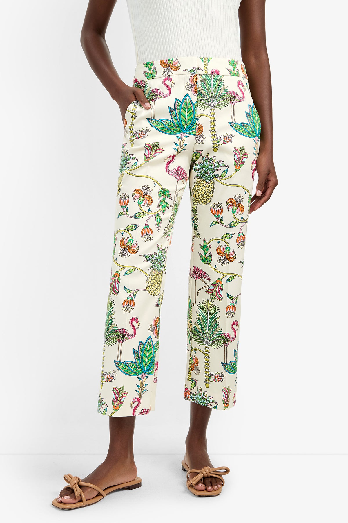 A person wears the LULU PANT, cream cotton-spandex pants with a tropical flamingo and floral print, paired with a white ribbed top and tan bow sandals.
