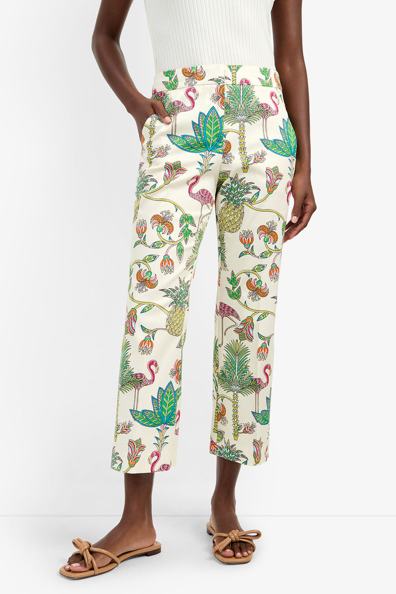 A person wears the LULU PANT, cream cotton-spandex pants with a tropical flamingo and floral print, paired with a white ribbed top and tan bow sandals.