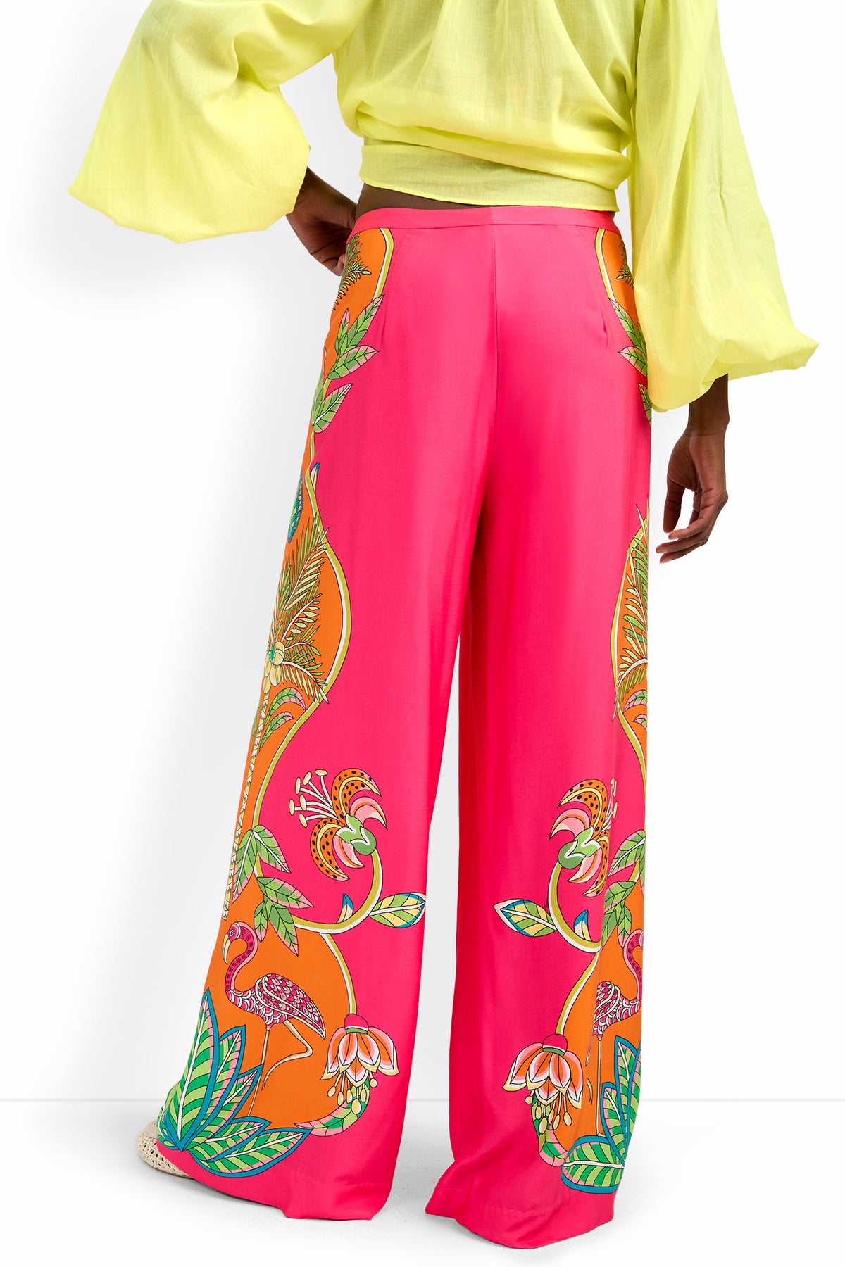 A person is seen from behind wearing the Lanay Pant—bright pink, wide-leg pants with colorful tropical bird and floral prints—and a loose, long-sleeve yellow top, set against a white background.