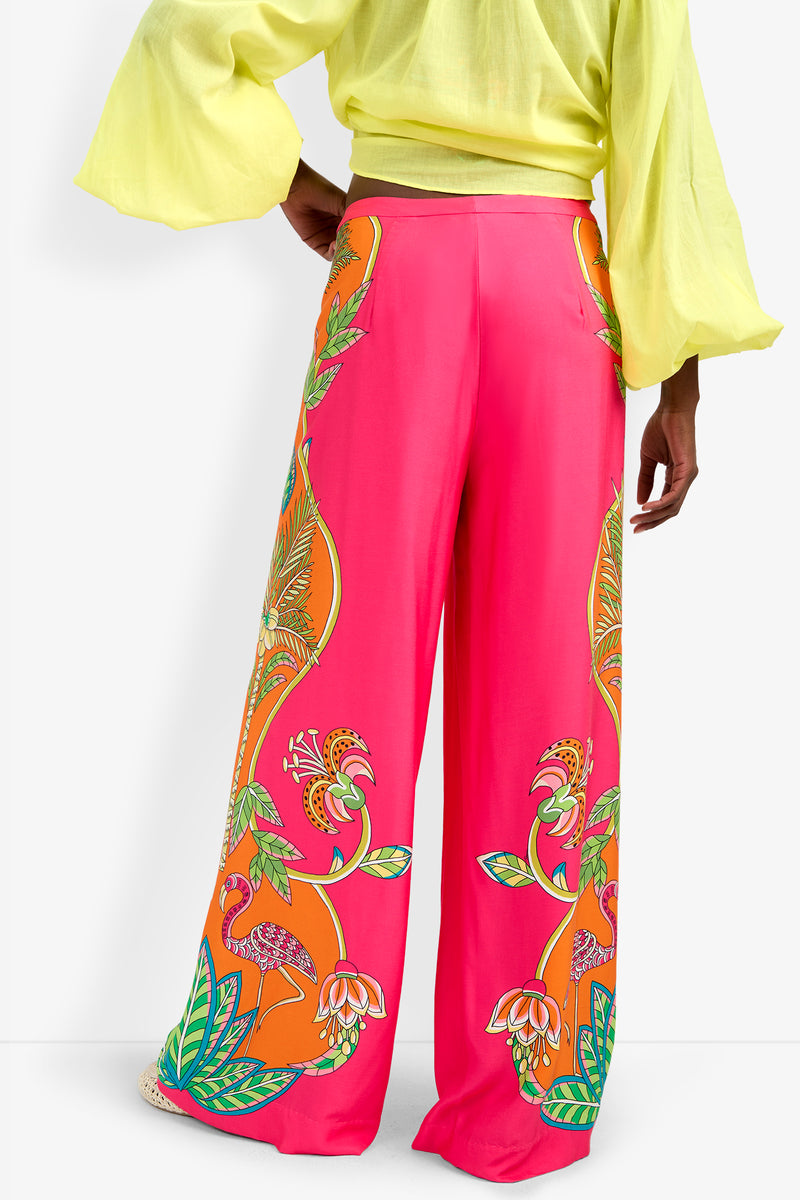 A person is shown from the back wearing the Lanay Pant in bright pink with tropical flamingo and flower designs, paired with a light yellow long-sleeve top featuring flared sleeves.