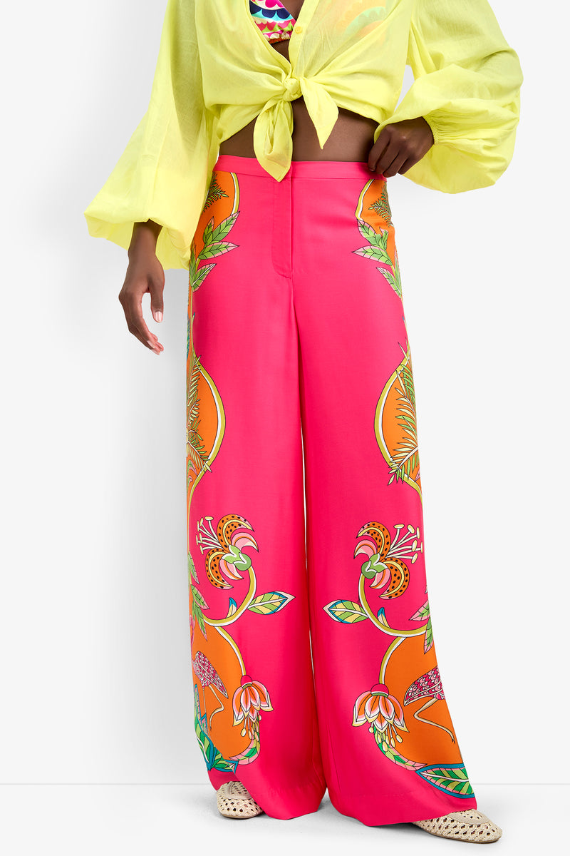 A person wears the Lanay Pant, featuring bright pink wide legs with bold orange and yellow floral patterns, paired with a yellow sheer blouse tied at the waist and woven sandals. Their head is not visible in the image.