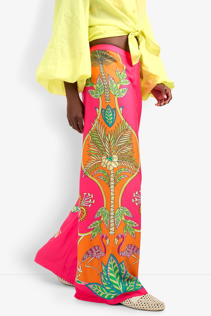 Someone wearing the Lanay Pant, featuring a vibrant tropical print in pink, orange, and green with a wide-leg fit, paired with a light yellow tie-front shirt and beige woven pointed shoes.