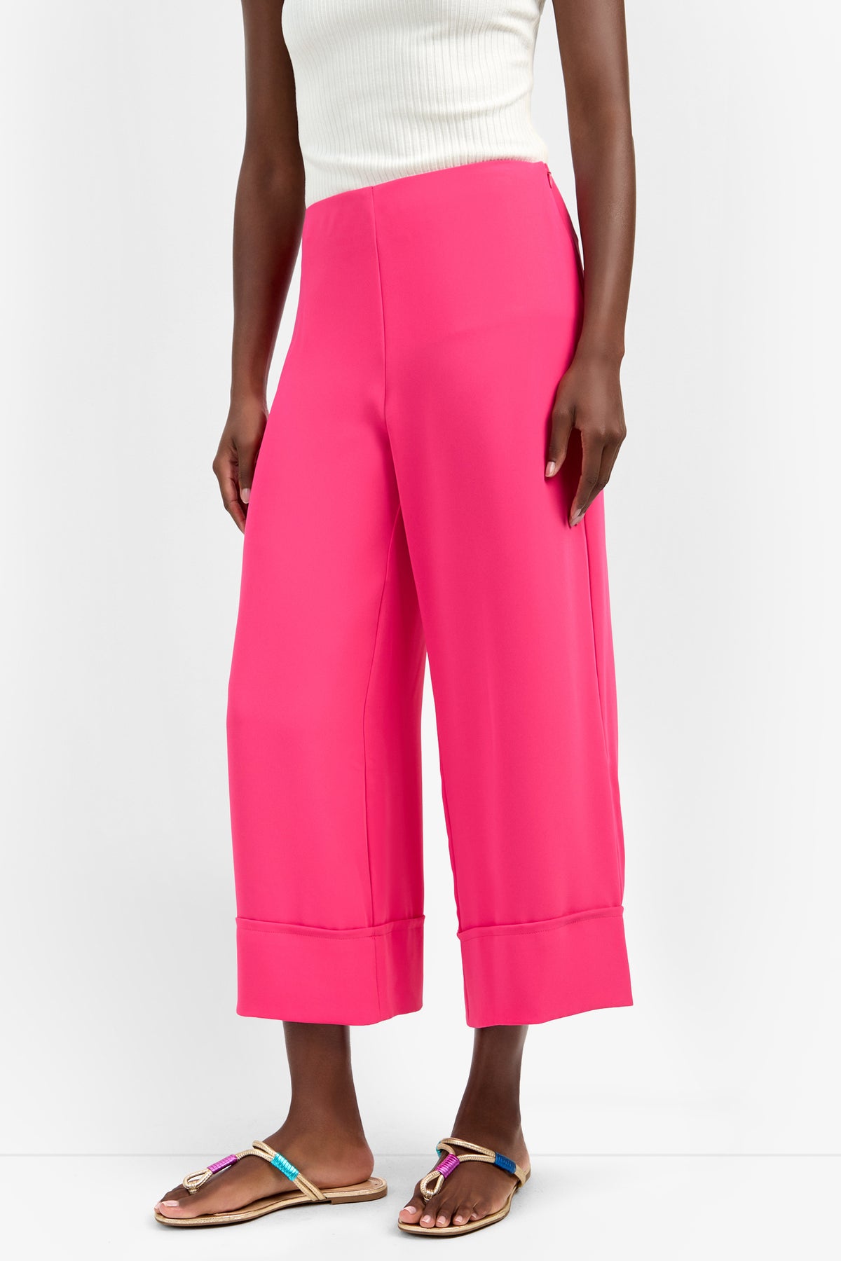 Wearing the bright pink BAHOOKA PANT—a wide-leg, cropped polyester style—paired with a white sleeveless ribbed top and colorful flat sandals, a person stands against a plain white background.