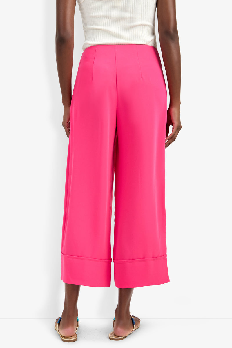 A person is seen from the back wearing the Bahooka Pant in bright pink, styled with a white sleeveless top and flat sandals, all set against a white background.