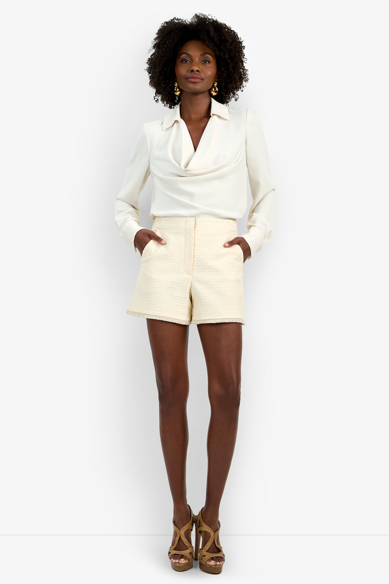 A woman stands with her hands in her pockets against a plain white background, wearing cream textured Valera Fringe Shorts, a white long-sleeve blouse, and brown high-heeled sandals.