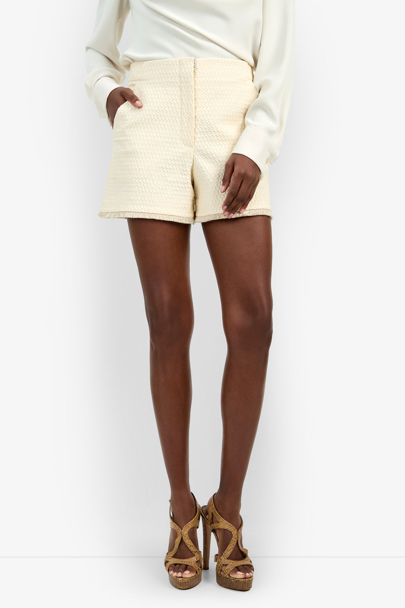 A woman stands with her hands in her pockets against a plain white background, wearing a cream blouse, the Valera Fringe Short in textured cream, and tan platform heels.
