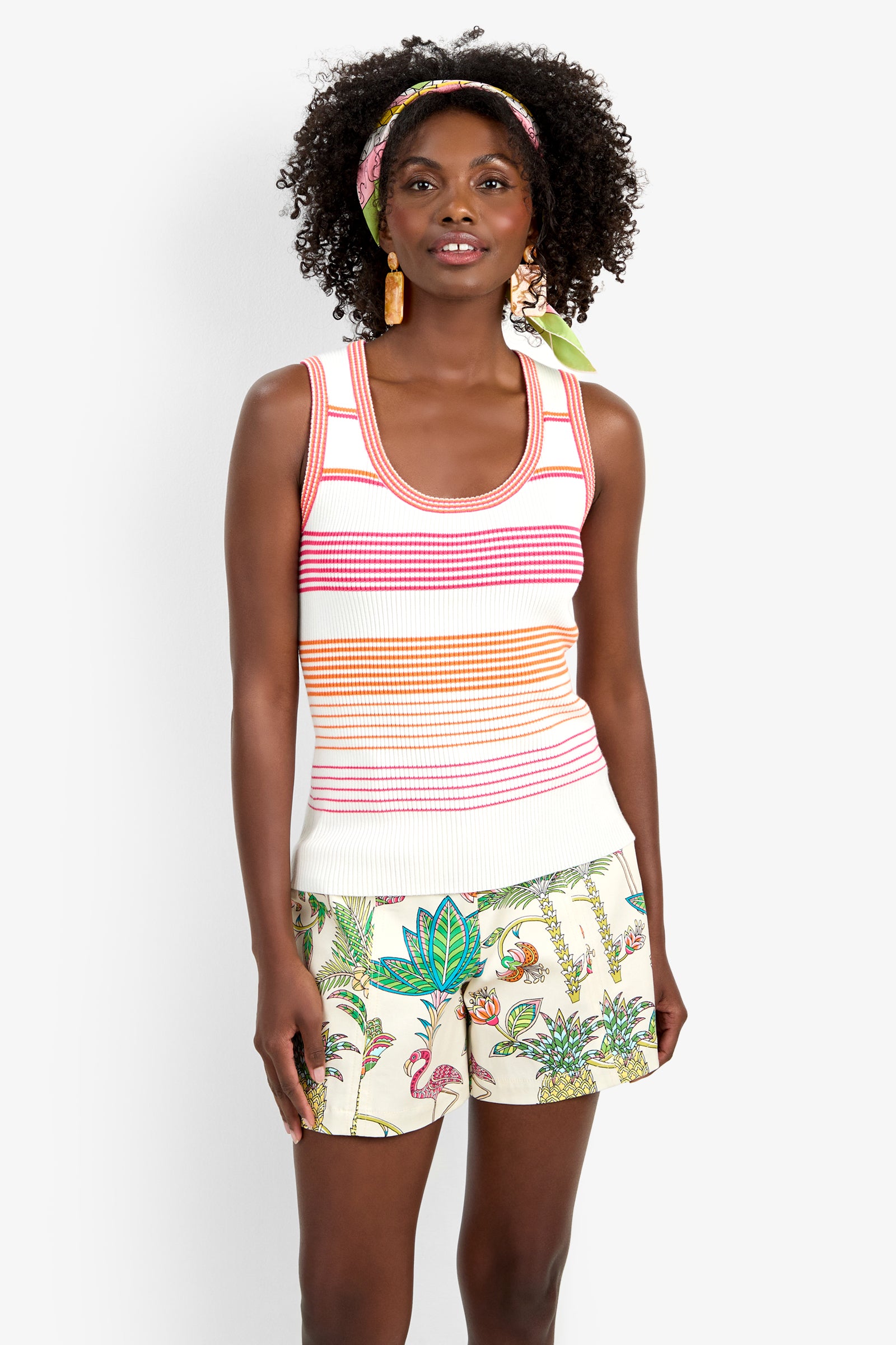 A woman with curly hair, a printed headband, and large earrings smiles while wearing the Paolini Tank and tropical print shorts featuring flamingos, plants, and animals.