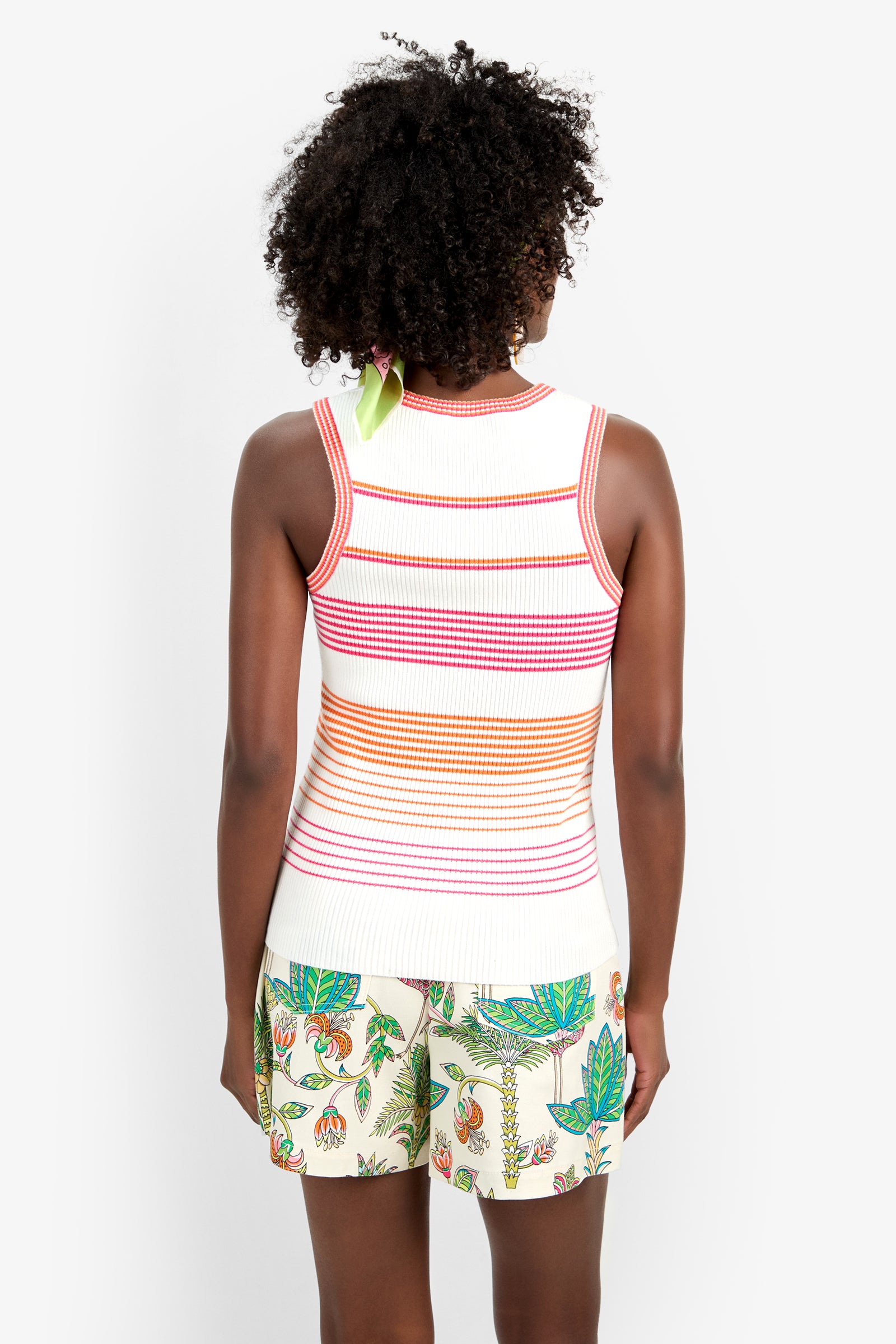 A person with curly hair stands facing away from the camera, wearing the Paolini Tank—a white sleeveless top with red and orange horizontal stripes—and white shorts featuring a colorful floral and fruit pattern.