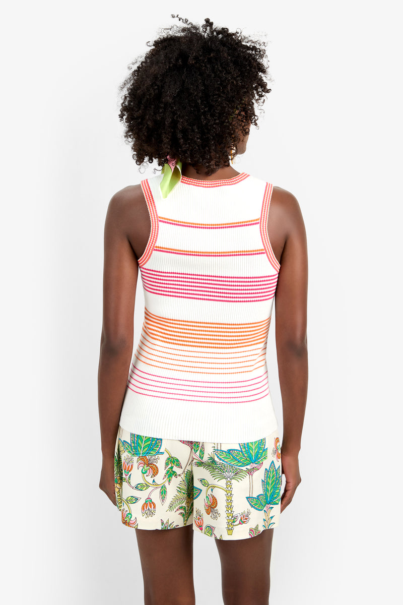A person with curly hair stands facing away from the camera, wearing the Paolini Tank—a white sleeveless top with red and orange horizontal stripes—and white shorts featuring a colorful floral and fruit pattern.