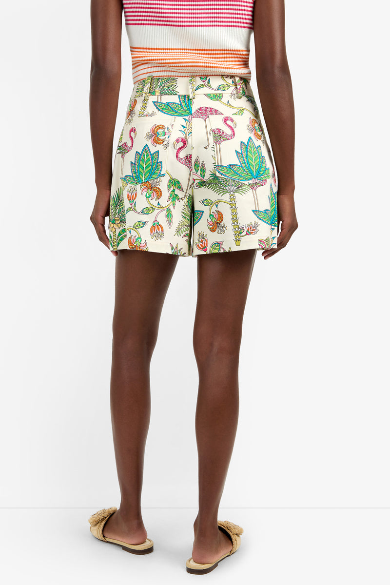 A person stands facing away, wearing the NORIAH SHORT—high-waisted cream cotton shorts with a colorful tropical print of flamingos, leaves, and pineapples—paired with a striped crop top and beige slide sandals.