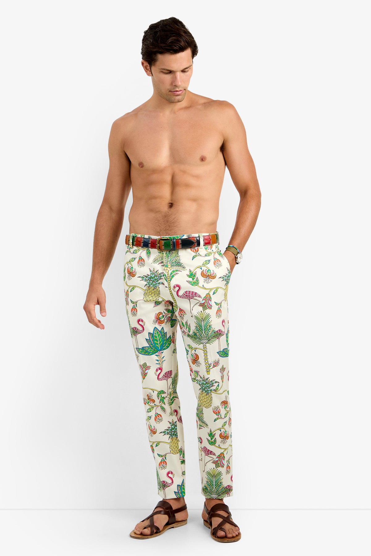 A shirtless man models the CLYDE SLIM TROUSER in MULTI, featuring vibrant tropical prints. He accessorizes with a plaid belt and brown sandals, standing against a plain white backdrop.