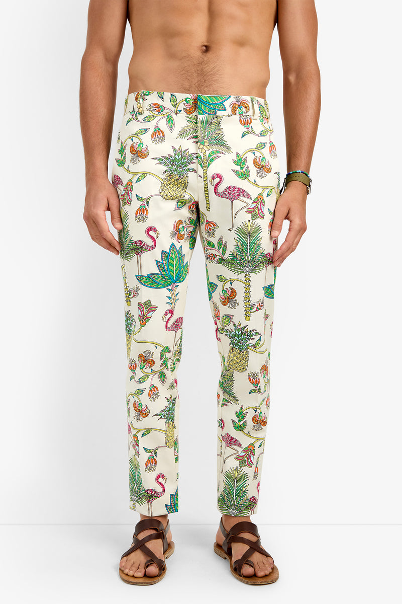 A shirtless person wears CLYDE SLIM TROUSER—cream cotton pants with a vibrant tropical print of flamingos, toucans, pineapples, and leaves—paired with brown sandals against a white background.