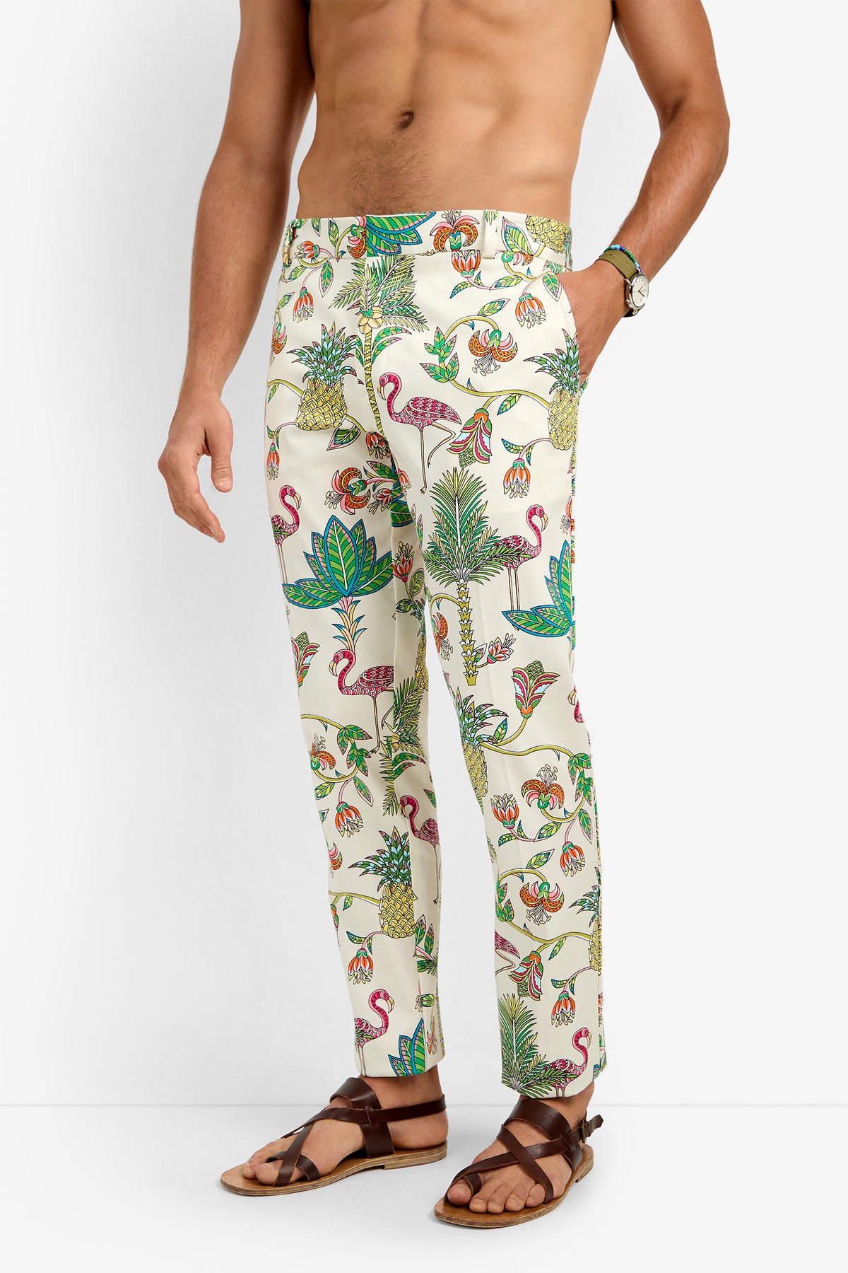 A shirtless man wears CLYDE SLIM TROUSER, cream-colored cotton lycra pants with a vibrant tropical print of flamingos, pineapples, and leaves. He pairs them with brown sandals and a wristwatch against a plain white background.