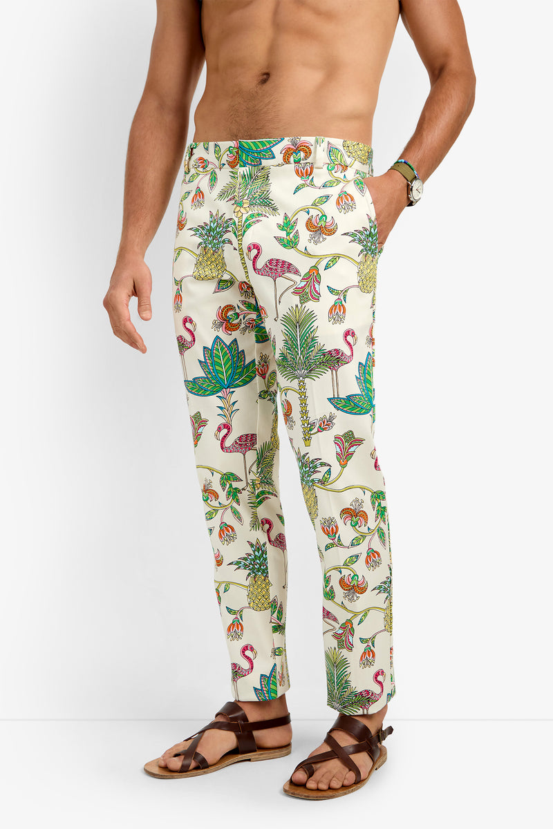 A shirtless man wears CLYDE SLIM TROUSER, cream-colored cotton lycra pants with a vibrant tropical print of flamingos, pineapples, and leaves. He pairs them with brown sandals and a wristwatch against a plain white background.