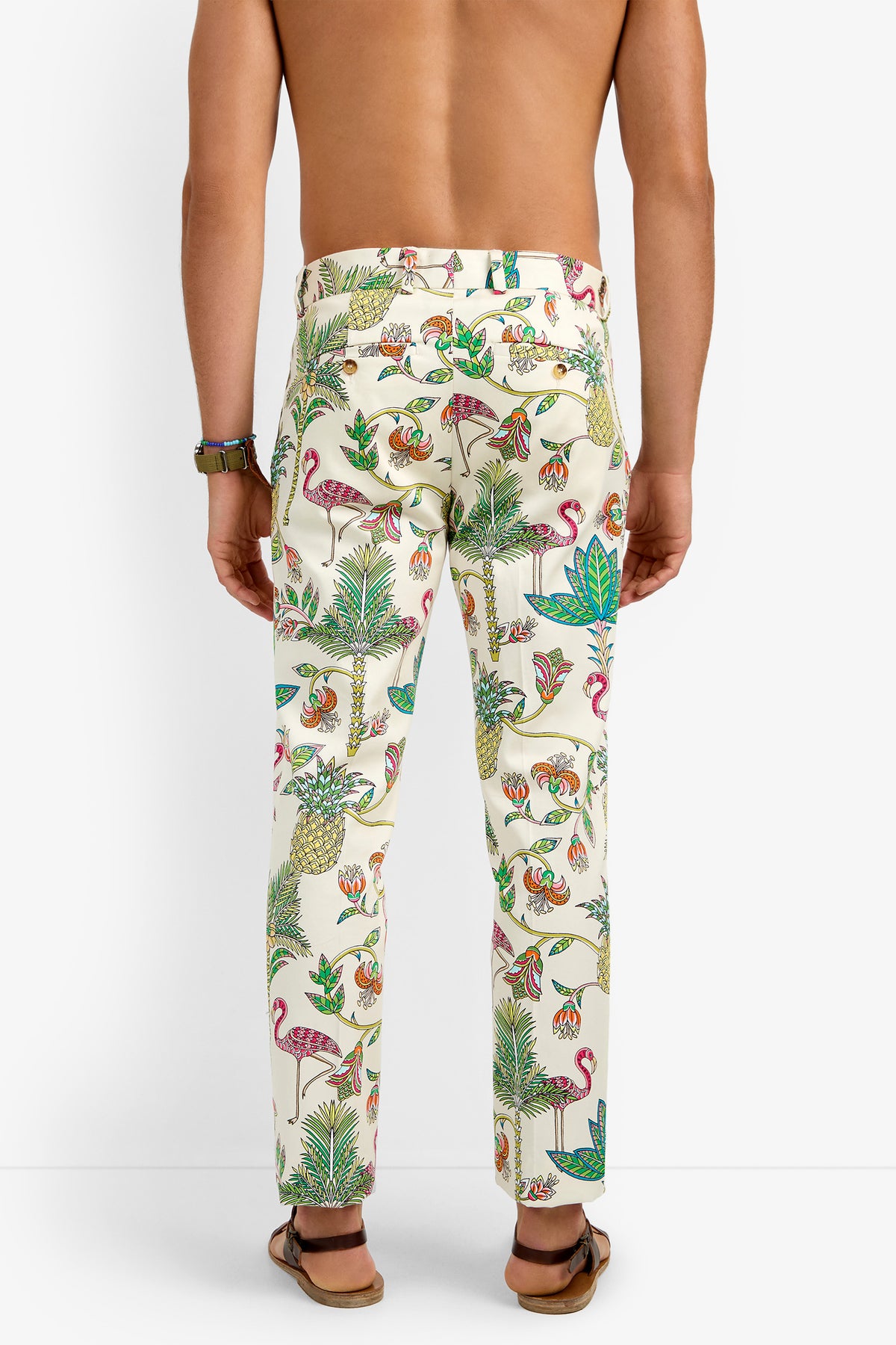 A person stands barefoot, facing away from the camera, wearing CLYDE SLIM TROUSER in a colorful tropical print with flamingos, plants, and birds; only their lower back and legs are visible.