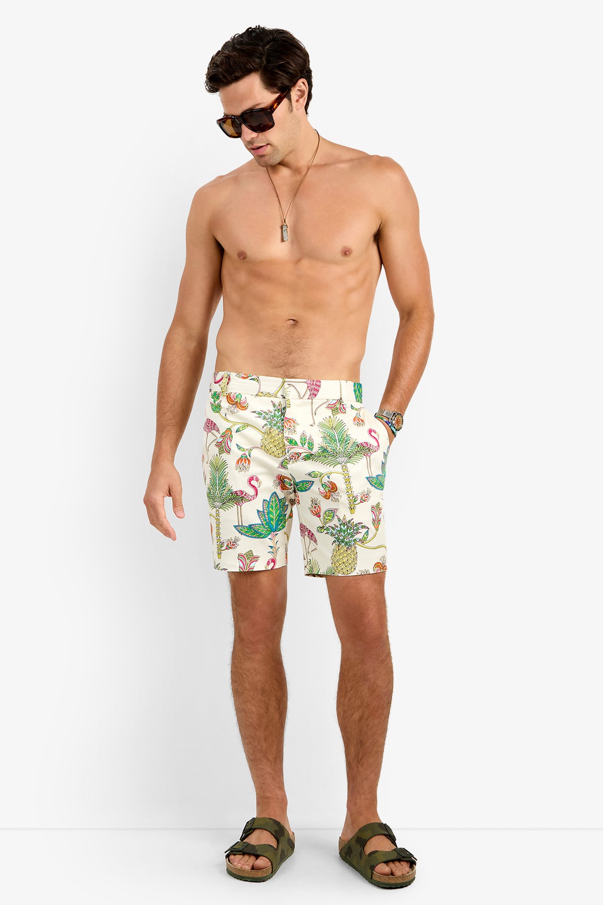 A shirtless man in sunglasses, a necklace, LAWRENCE SHORT in MULTI, and green sandals stands looking down against a plain white background.