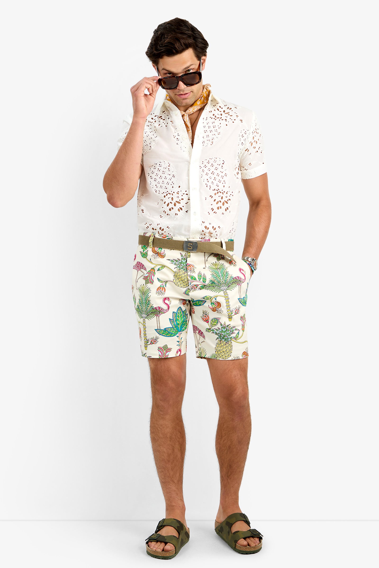 A man stands against a white background wearing sunglasses, a patterned scarf, a white cotton lycra eyelet shirt, the LAWRENCE SHORT in tropical print, olive green sandals, and a brown belt.