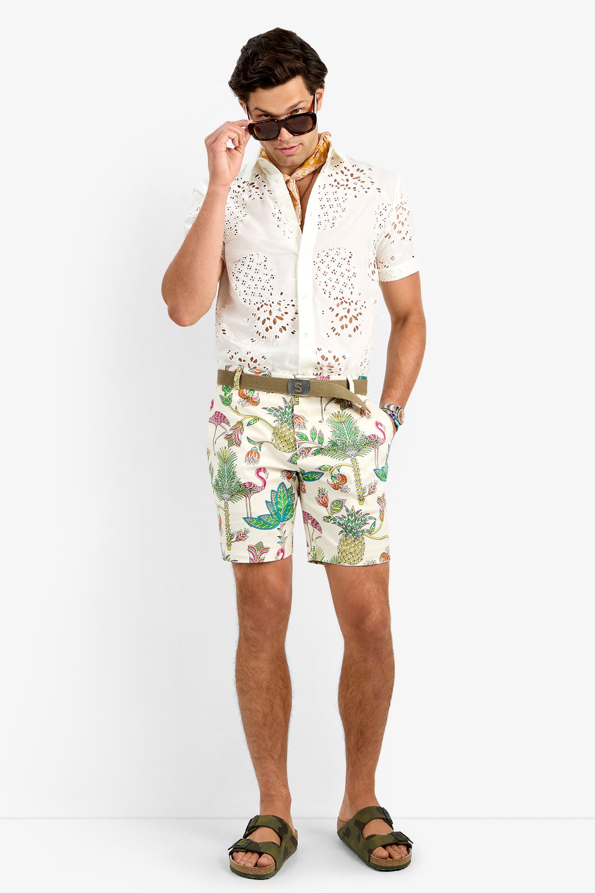A man stands against a white background wearing sunglasses, a patterned scarf, a white cotton lycra eyelet shirt, the LAWRENCE SHORT in tropical print, olive green sandals, and a brown belt.