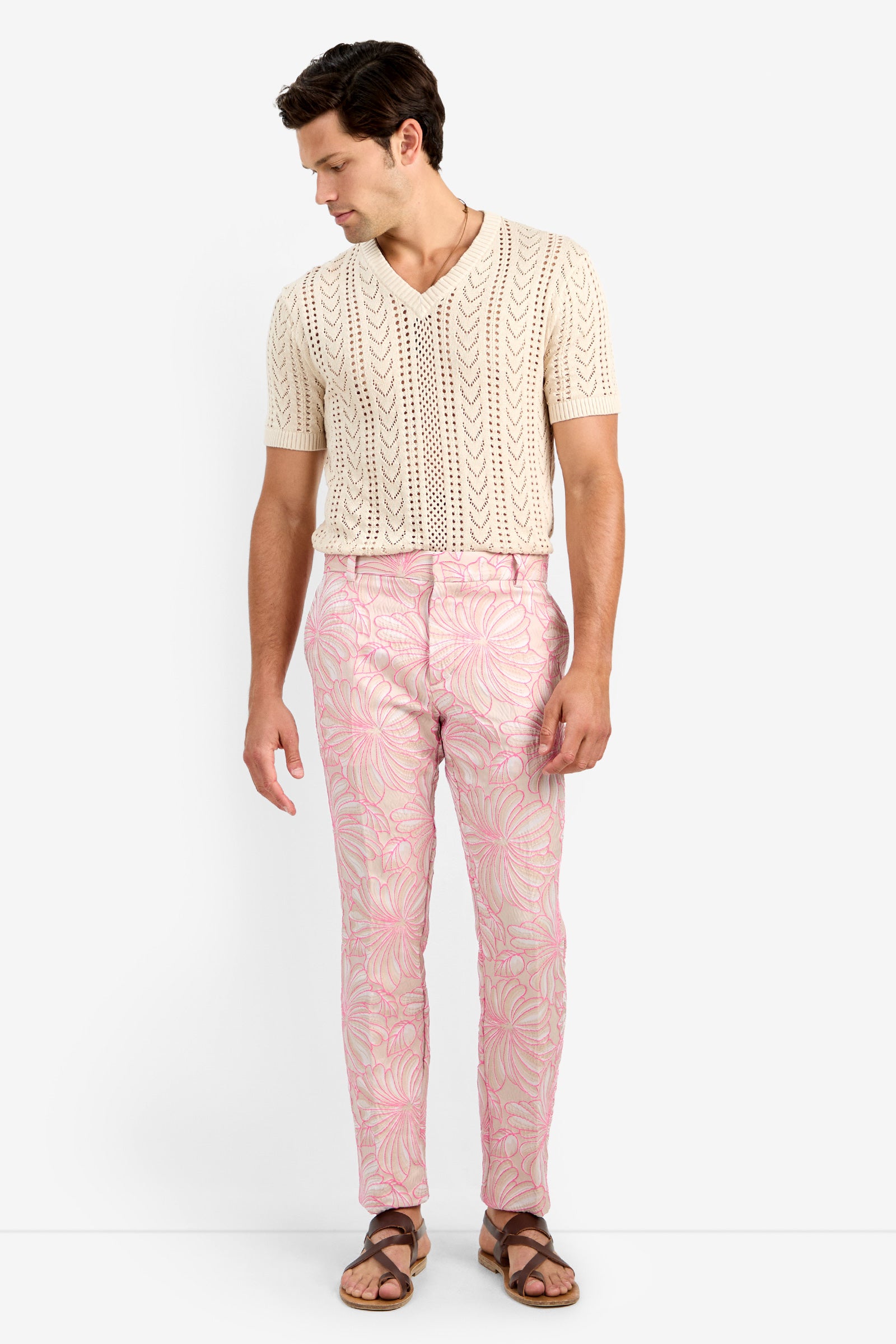 A man stands against a white background, wearing pink leafy-patterned CLYDE SLIM TROUSER made from lycra, a cream cotton short-sleeve knit shirt, and brown sandals. He looks down to his left with arms relaxed at his sides.