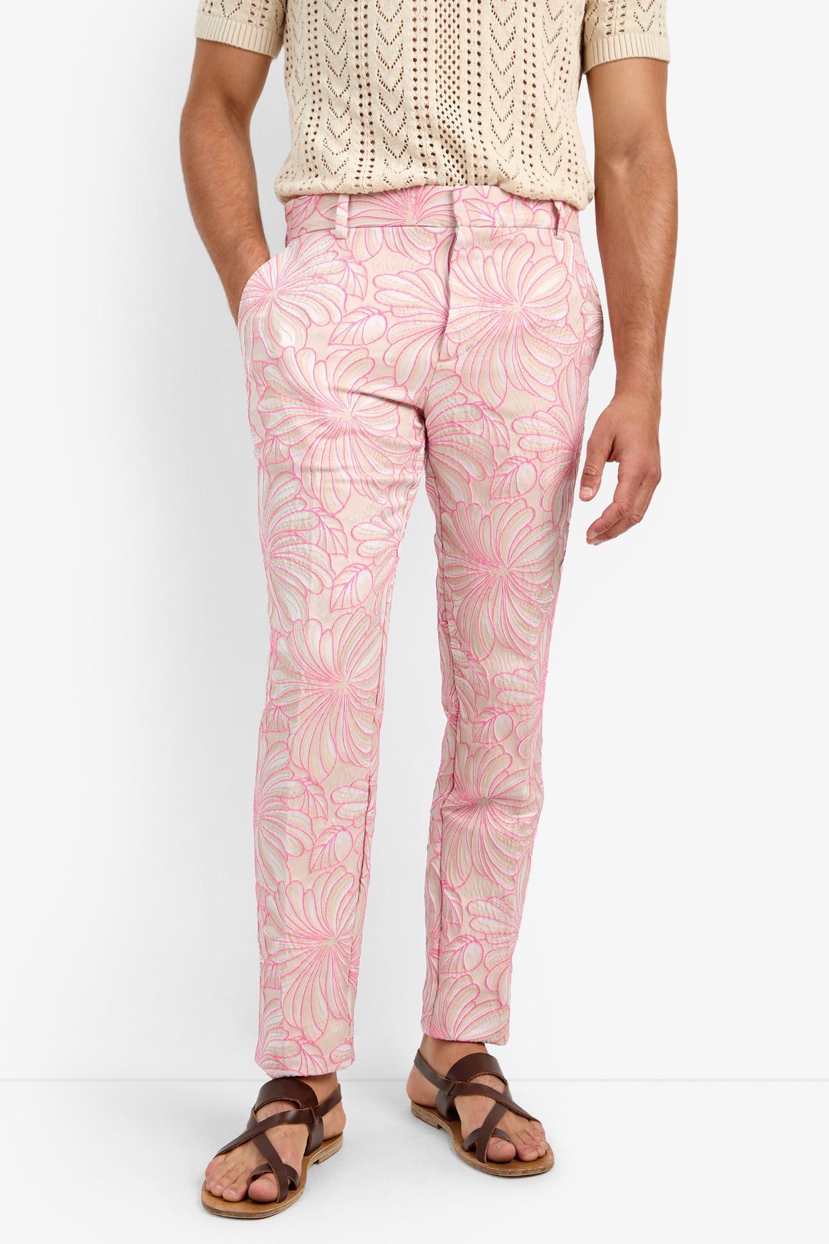 Wearing the CLYDE SLIM TROUSER in light pink cotton with a white and dark pink floral print, paired with a beige short-sleeve knit shirt and brown sandals, against a white background.