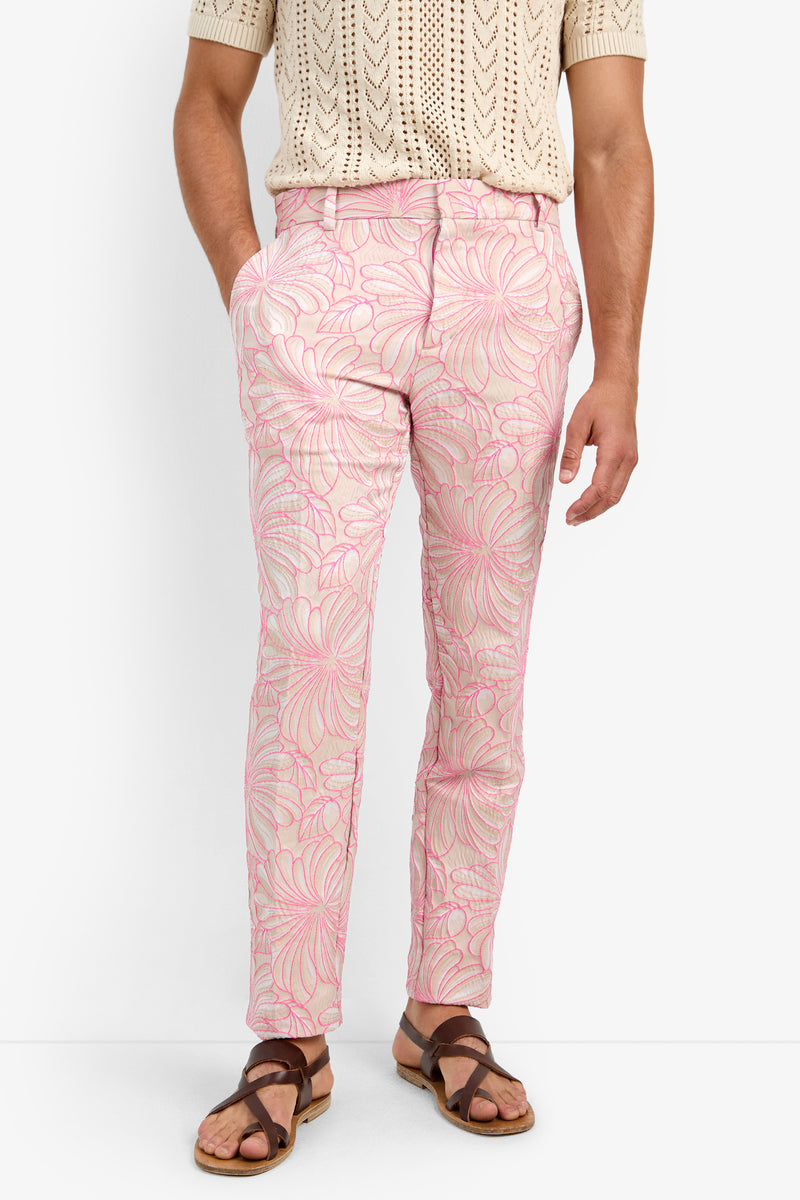 Wearing the CLYDE SLIM TROUSER in light pink cotton with a white and dark pink floral print, paired with a beige short-sleeve knit shirt and brown sandals, against a white background.