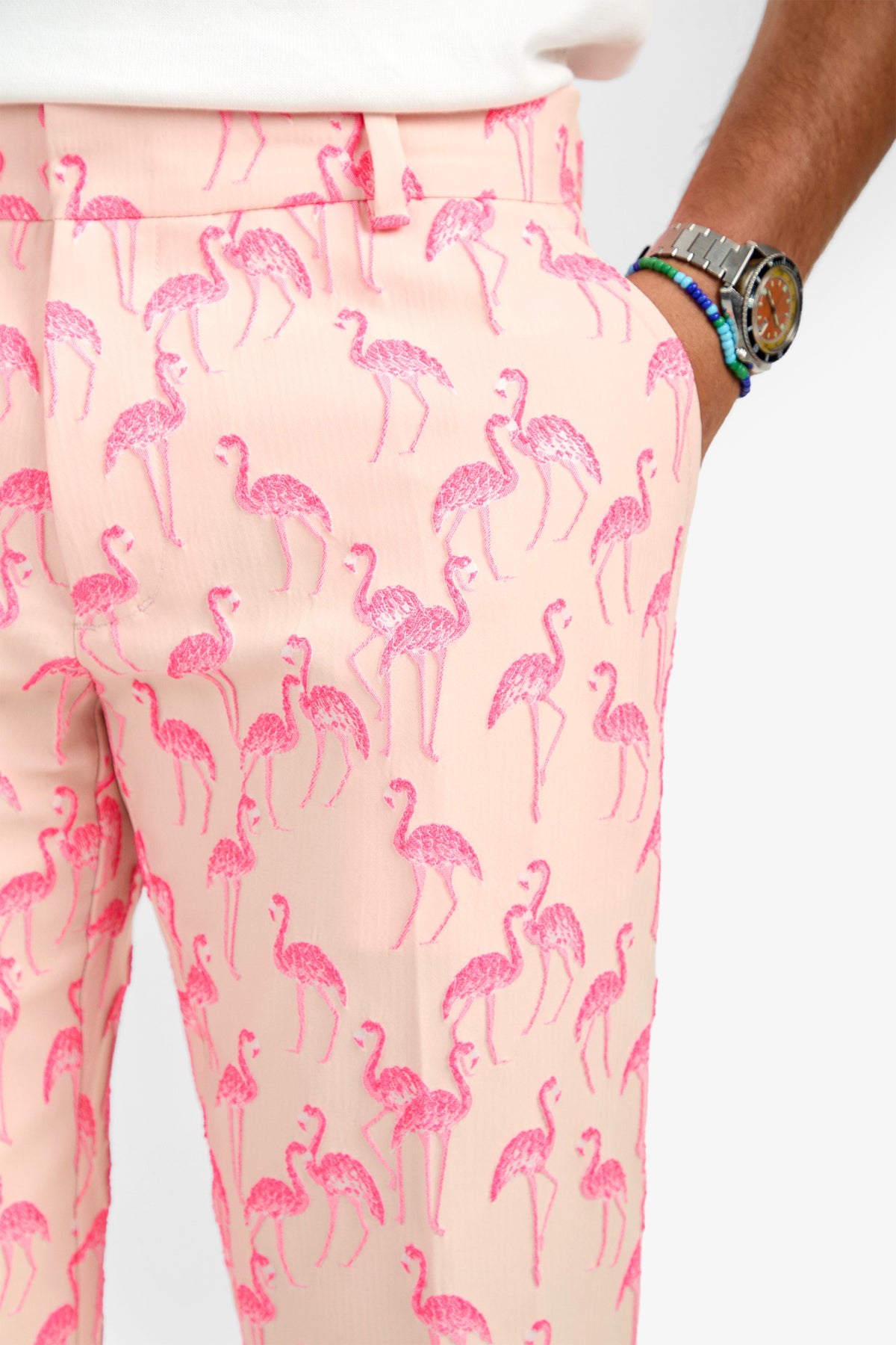 A person wearing the CLYDE SLIM TROUSER in light pink with bright pink flamingos, made of soft polyester, paired with a tucked-in white shirt and a wristwatch with colorful beaded bracelets.