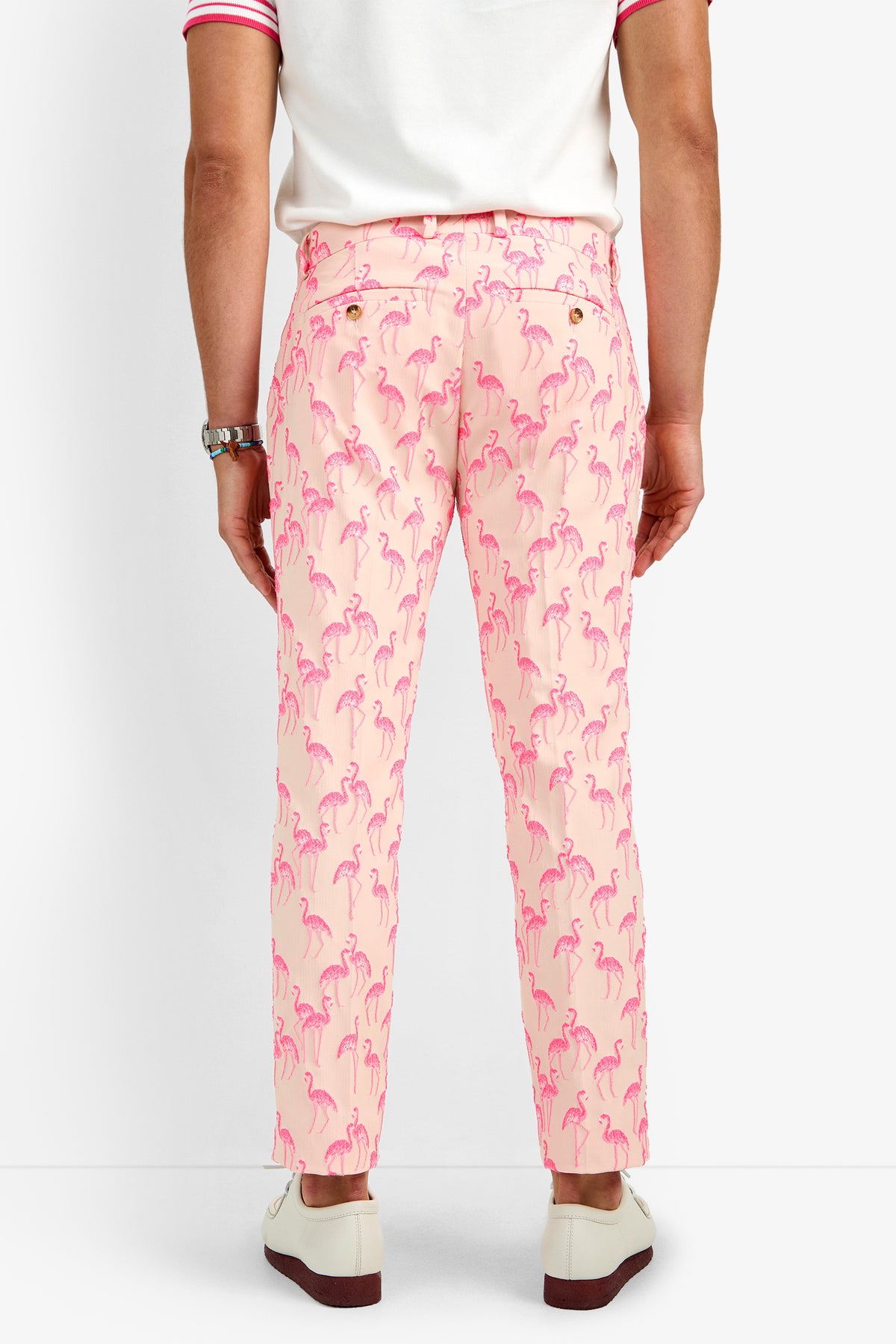 A person faces away from the camera, dressed in CLYDE SLIM TROUSER—light pink pants with bright pink flamingo prints made of soft polyamide—paired with a white shirt featuring pink trim and white loafers.