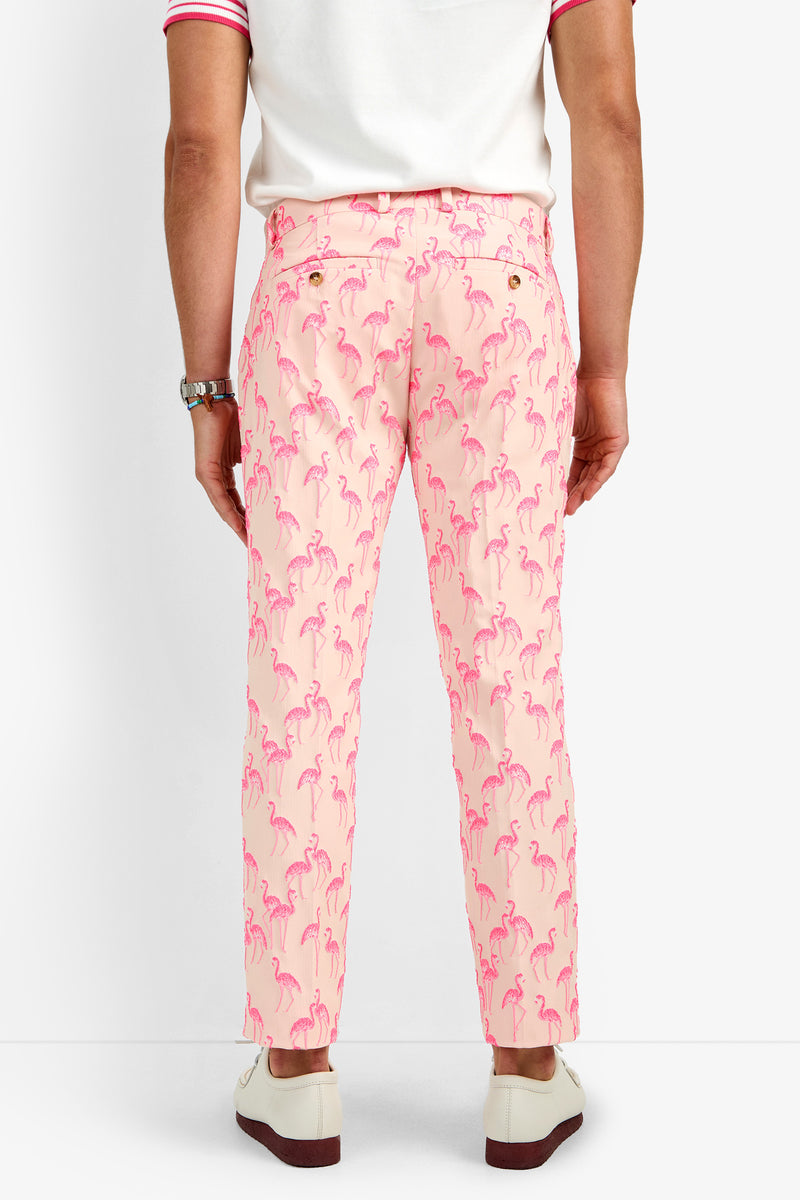 A person faces away from the camera, dressed in CLYDE SLIM TROUSER—light pink pants with bright pink flamingo prints made of soft polyamide—paired with a white shirt featuring pink trim and white loafers.