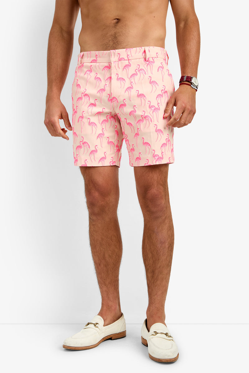 A man wears light pink LAWRENCE SHORTS with a pink flamingo print, a brown watch, and cream loafers. He is shirtless and standing against a white background.