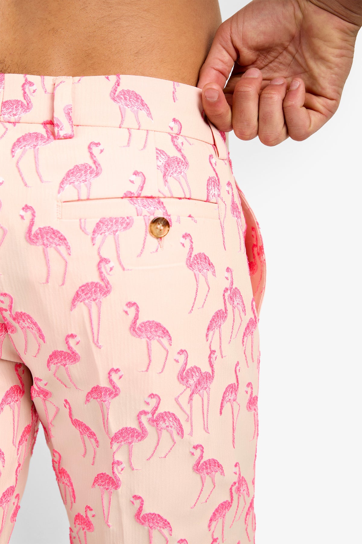 Close-up of a person wearing LAWRENCE SHORT, light pink polyester shorts with pink flamingos, pulling the waistband away from their waist. A back pocket with a button is visible.